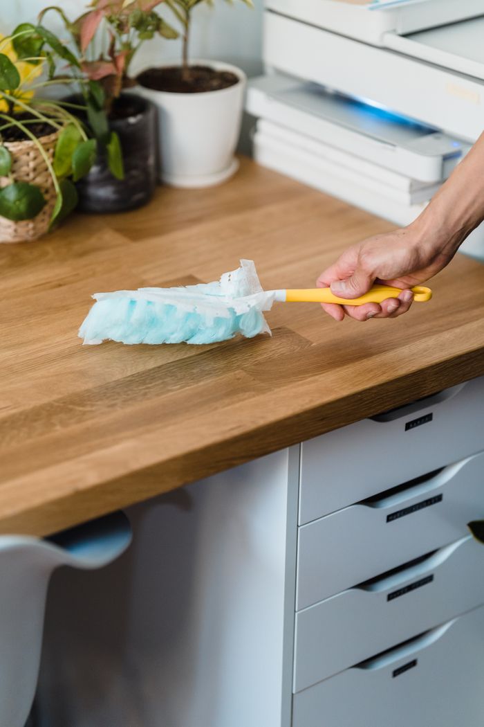 about-us-02 A hand using a feather duster to clean a wooden desk, surrounded by plants and office items.